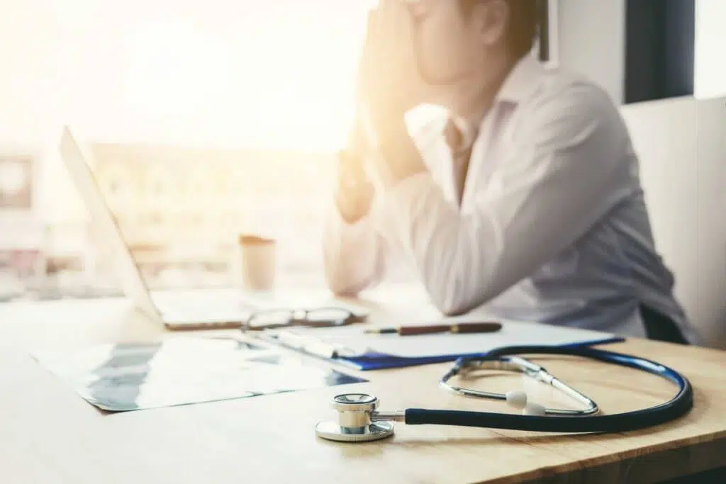tired physician shown blurred in the background at desk covering face
