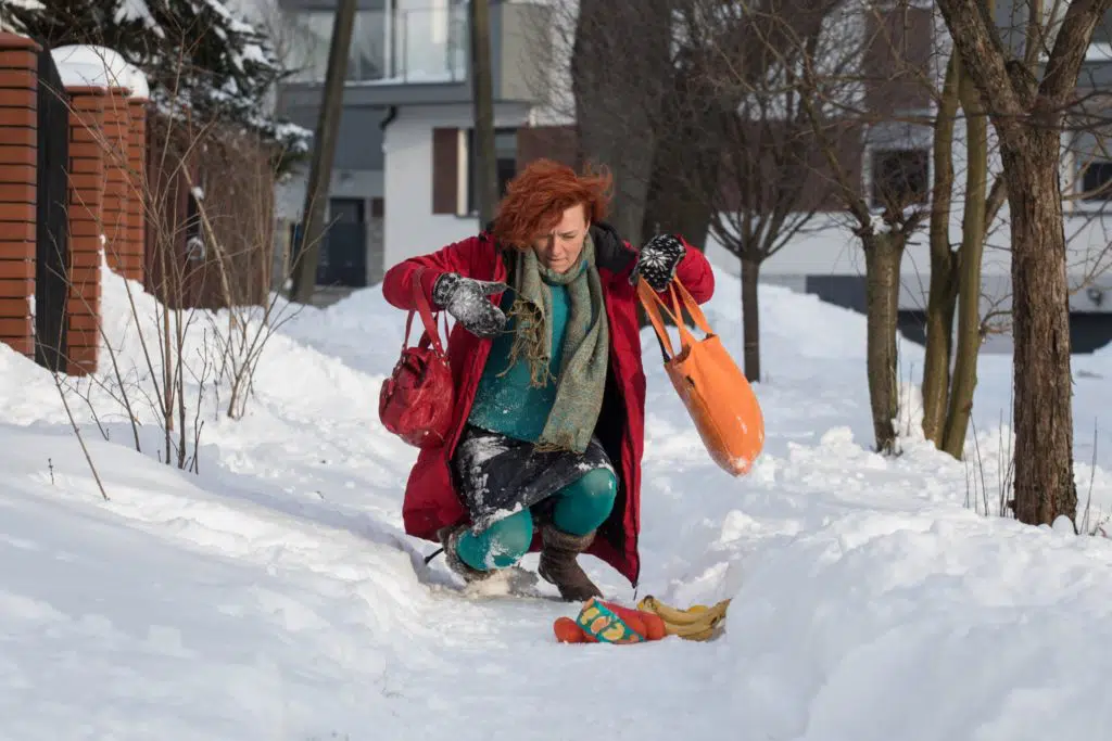 Woman with red hair and blue snow suit mid-fall on a snowy sidewalk