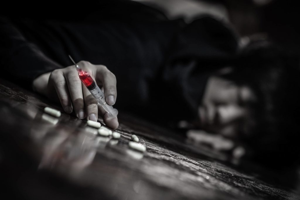 hand of a person laying on the floor holding a syringe with red fluid and surrounded by white pills