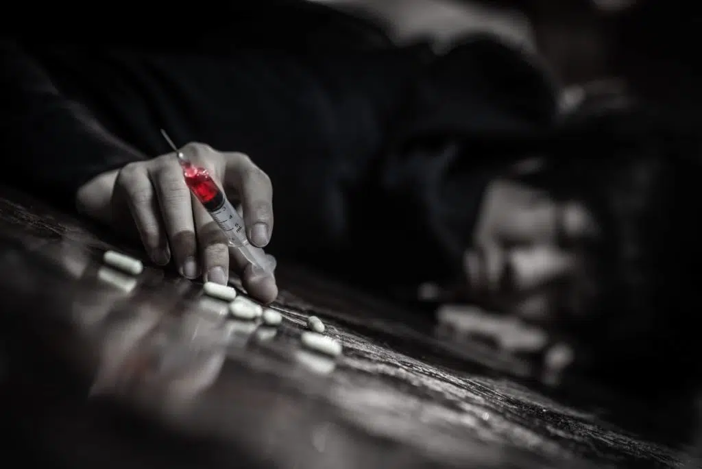 hand of a person laying on the floor holding a syringe with red fluid and surrounded by white pills