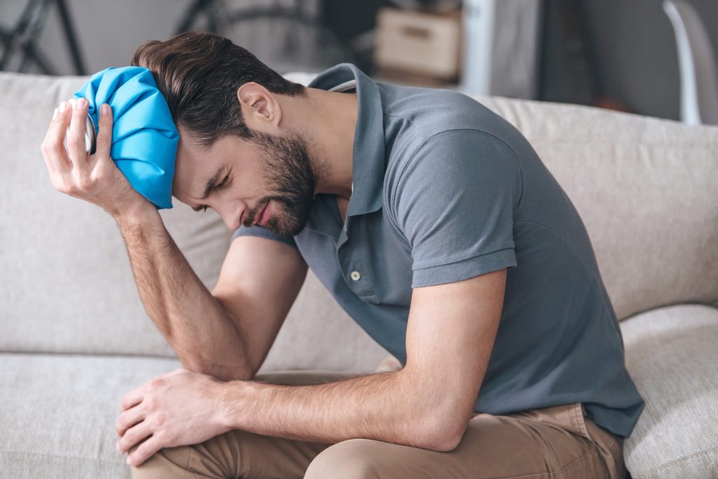 Young man with brown hair holds an ice pack on his head and looks in pain.