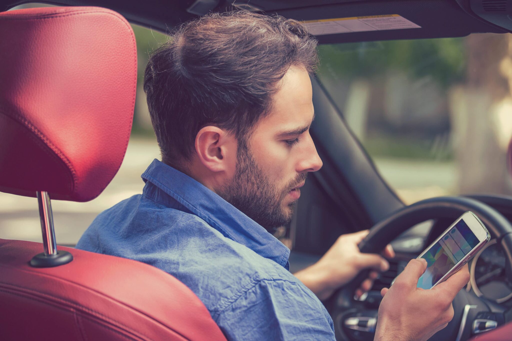 Man in the driver's seat of a car looks at his smartphone while driving.