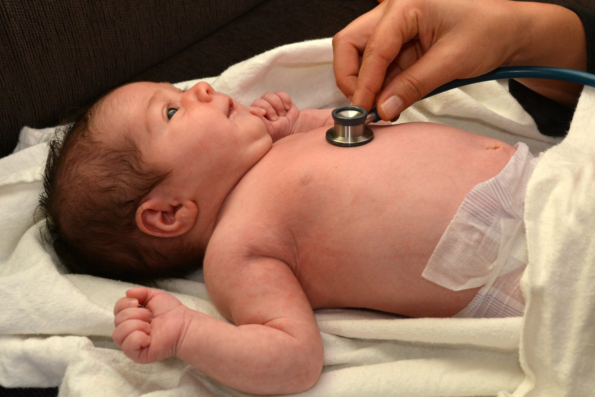 smiling baby in a diaper on a white blanket with a person's hand holding a stethoscope to their heart