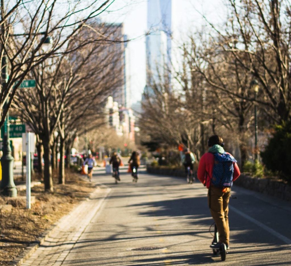 Person rides on a scooter in a NYC park toward the 1 World Trade Center building shown in the background.