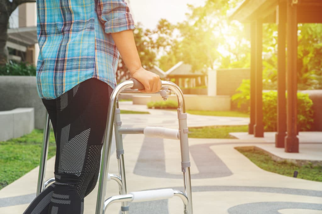 A person in a plaid shirt and athletic pants walks with an aluminum walker toward a setting sun in an outdoor courtyard.