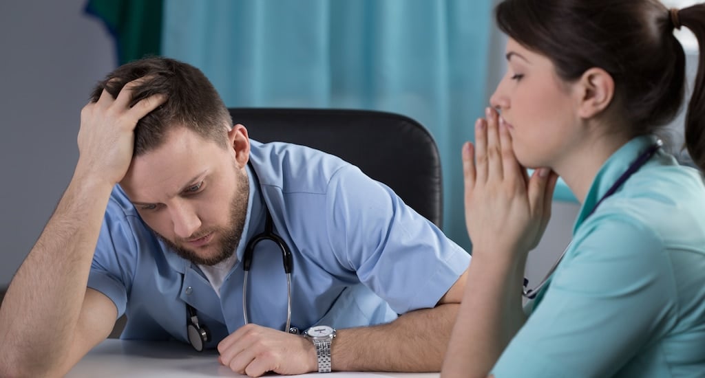 Young woman doctor closes eyes with hands to her mouth, while a young male doctor sitting next to her looks preoccupied after they both realize a medical mistake