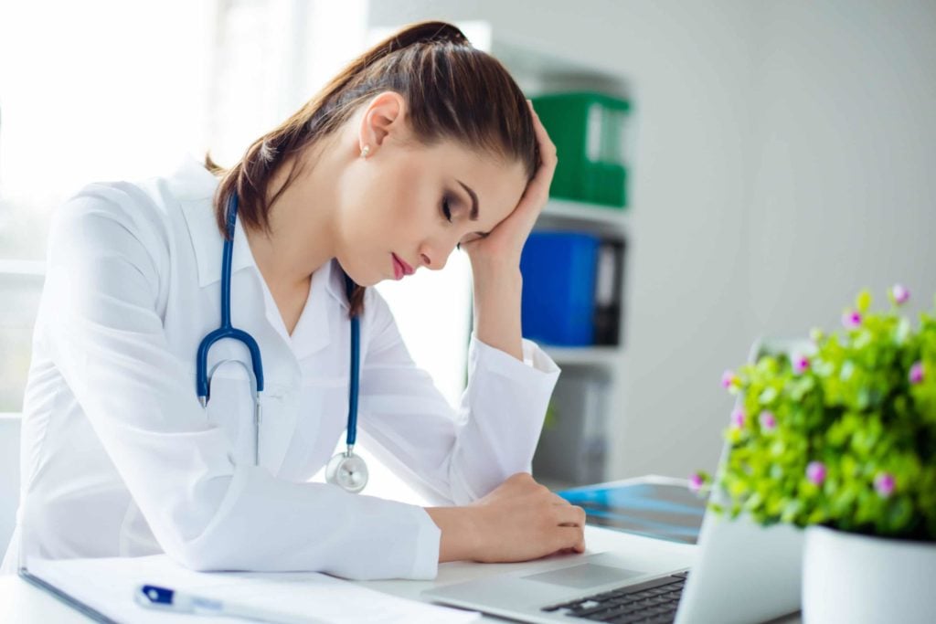Young physician with brown hair closes eyes at desk, resting her head in her hand since she is tired.