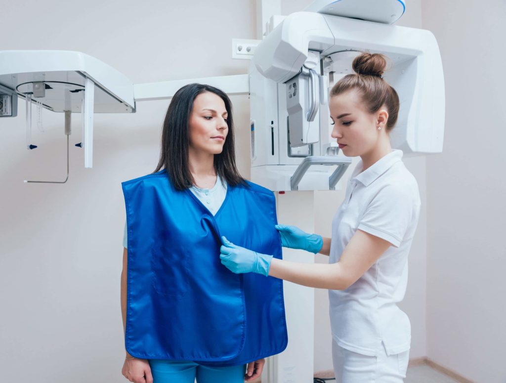 Woman wearing a protective mask around her shoulders is helped by a medical technician before an x-ray breast cancer screening.