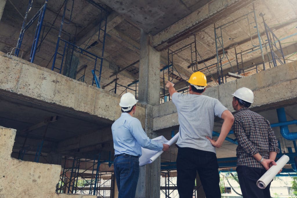 Three men in construction hard hats stand at a construction job site. One in the middle in a yellow safety helmet point up at the second level on the site.