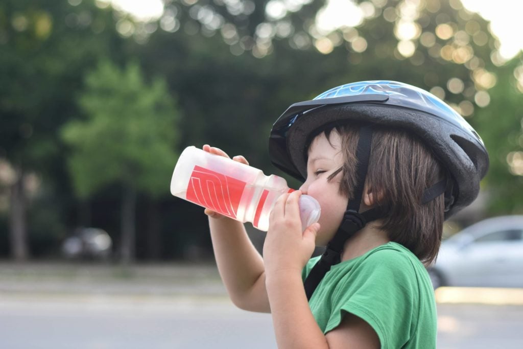 Child wearing a bike helmet and green shirt drinks from a water bottle