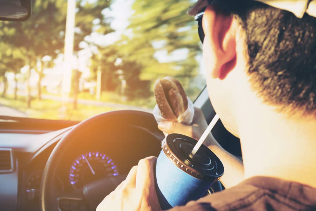 from behind the dashboard of a vehicle, a man drives distracted while eating a burger and drinking a soft drink