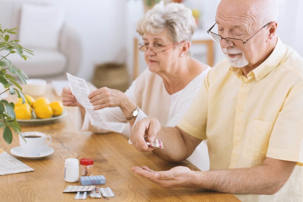 An elderly couple sits at a table. One takes medication while the other reads documentation.