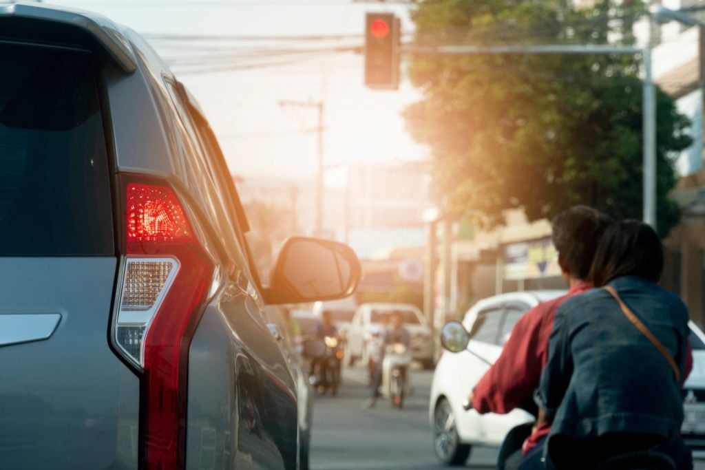 A car and motorbike wait at a red light at a busy intersection with the sun setting in the background.