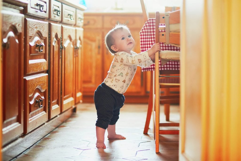 A baby stands herself up by holding onto a chair in a light-filled kitchen