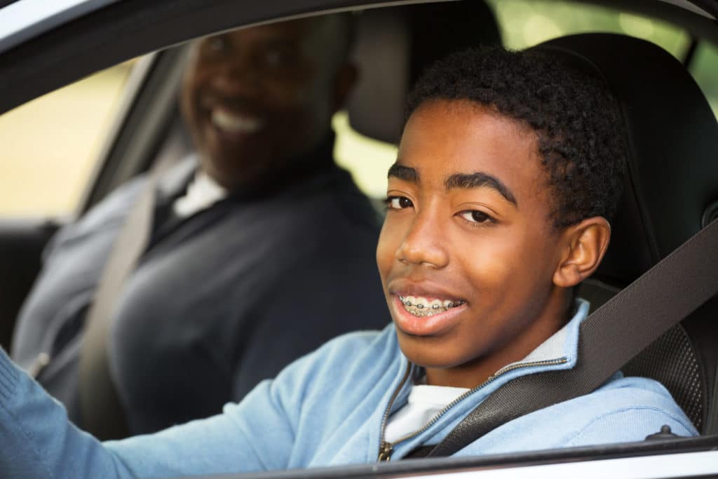 young teen driver sits behind the wheel with an adult smiling in the passenger seat.