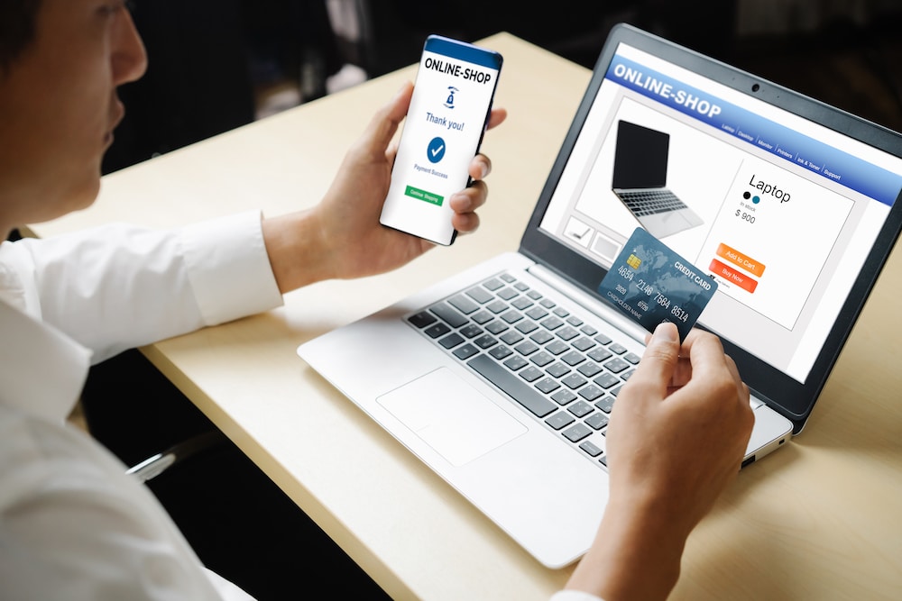 Young man looks at his phone and compares his credit card to the computer screen as he purchases a product.