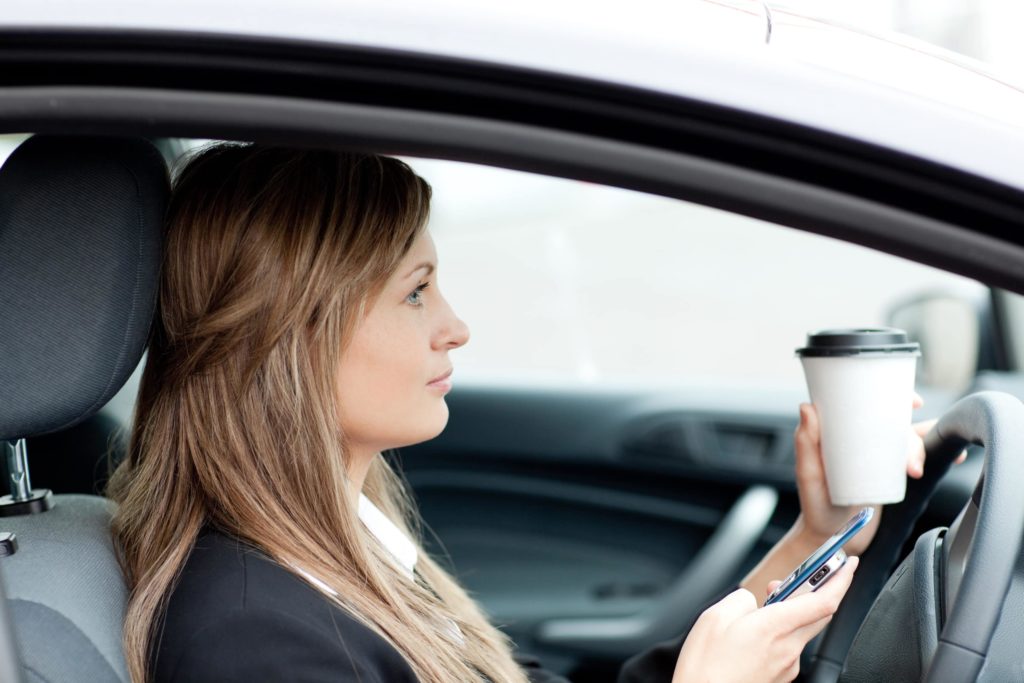 Woman in black suit jacket sits distracted behind the wheel in the driver seat with her phone and a coffee in her hands.