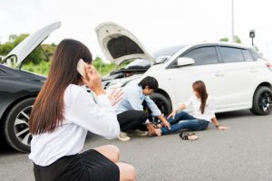 An Asian woman, feeling stressed, sits by the roadside following a traffic accident, experiencing the aftermath of a wreck and the strain it brings.