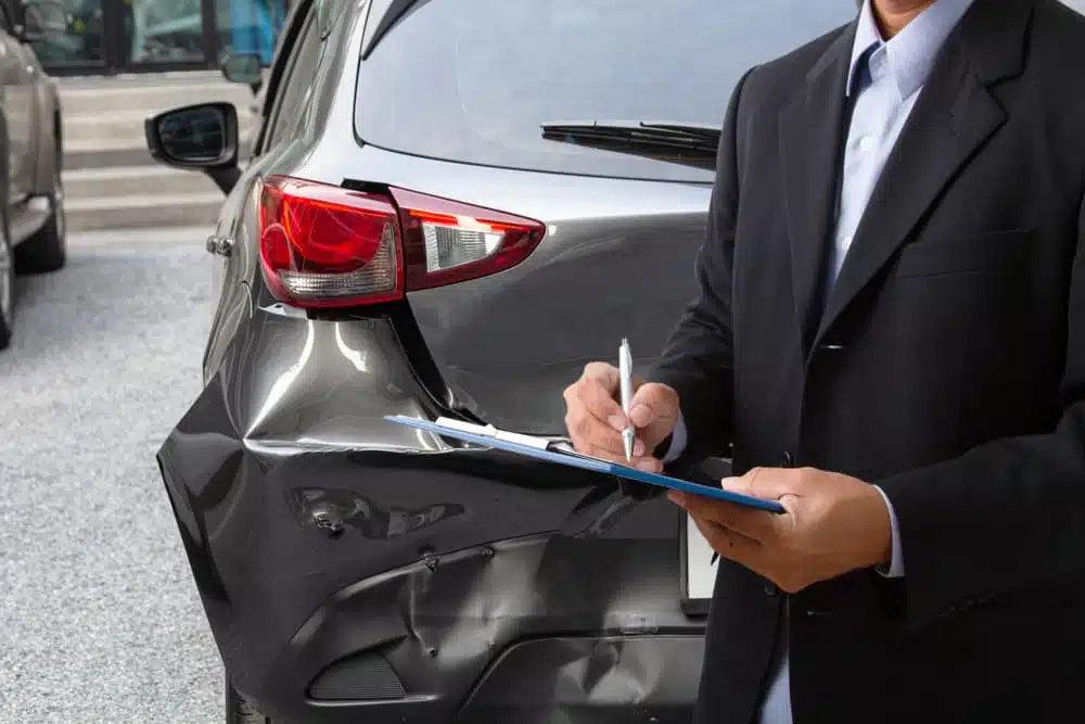 Insurance adjuster inspecting damage on a car's rear end, taking notes on a clipboard.