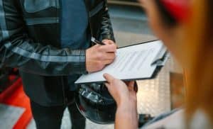 Motorcyclist in leather jacket signing documents, held by another person, with helmet in hand.