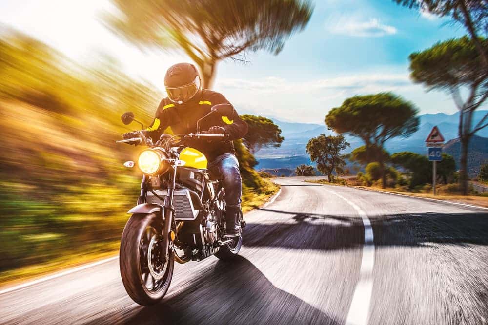 Motorcyclist in full gear riding fast on a curvy road with scenic mountain backdrop.