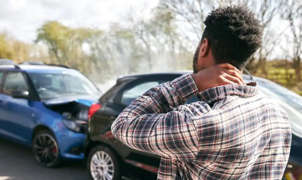 Young man rubbing neck in pain, standing by damaged car after traffic accident.