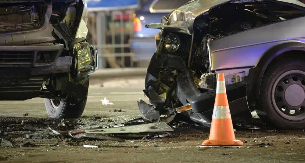 Two severely damaged cars in a nighttime accident scene with debris and a traffic cone.