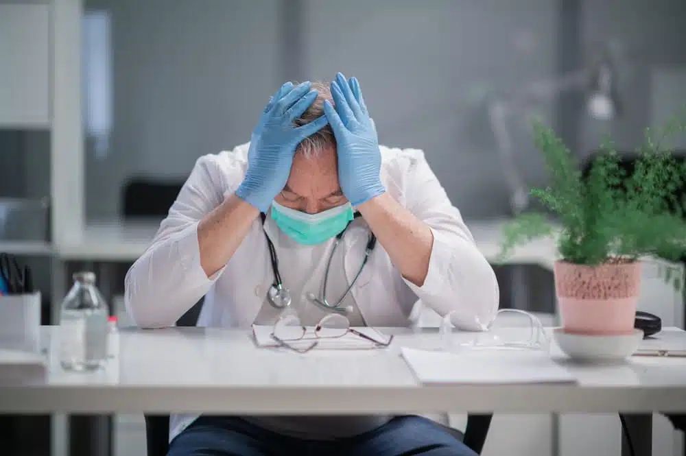 A distressed doctor wearing a mask and gloves sits at a desk.