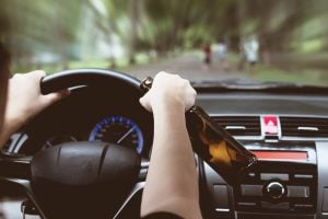 Woman drinking a beer while driving a car, illustrating the dangers of drunk driving.