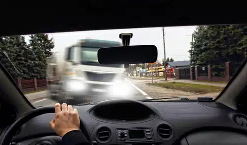 A driver’s view from inside a car shows their hand on the steering wheel, with an oncoming large white truck approaching at high speed on a two-lane road. The truck's headlights are blurred, indicating rapid movement, creating a potentially dangerous situation.