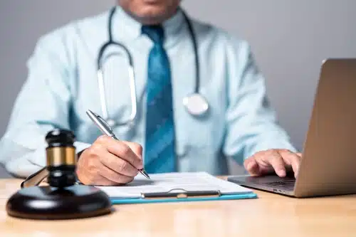 A medical professional, with a stethoscope around their neck, writing on a document while using a laptop. A gavel is placed on the desk, symbolizing a connection to medical law or healthcare compliance