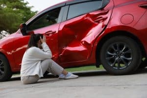 Woman sitting beside a red car with severe side damage.