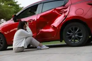 Woman sitting beside a red car with severe side damage.