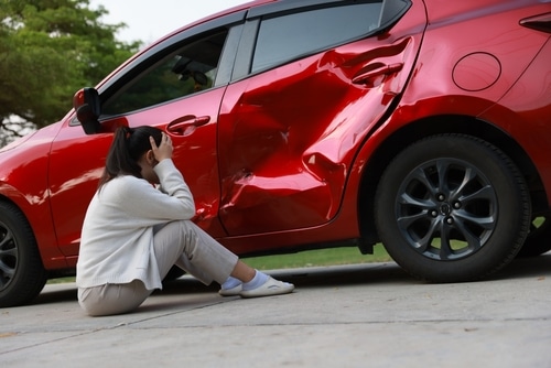 Woman sitting beside a red car with severe side damage.