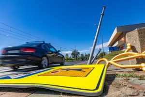 Car driving past a fallen pedestrian crossing sign and damaged pole after a traffic accident on a sunny day.